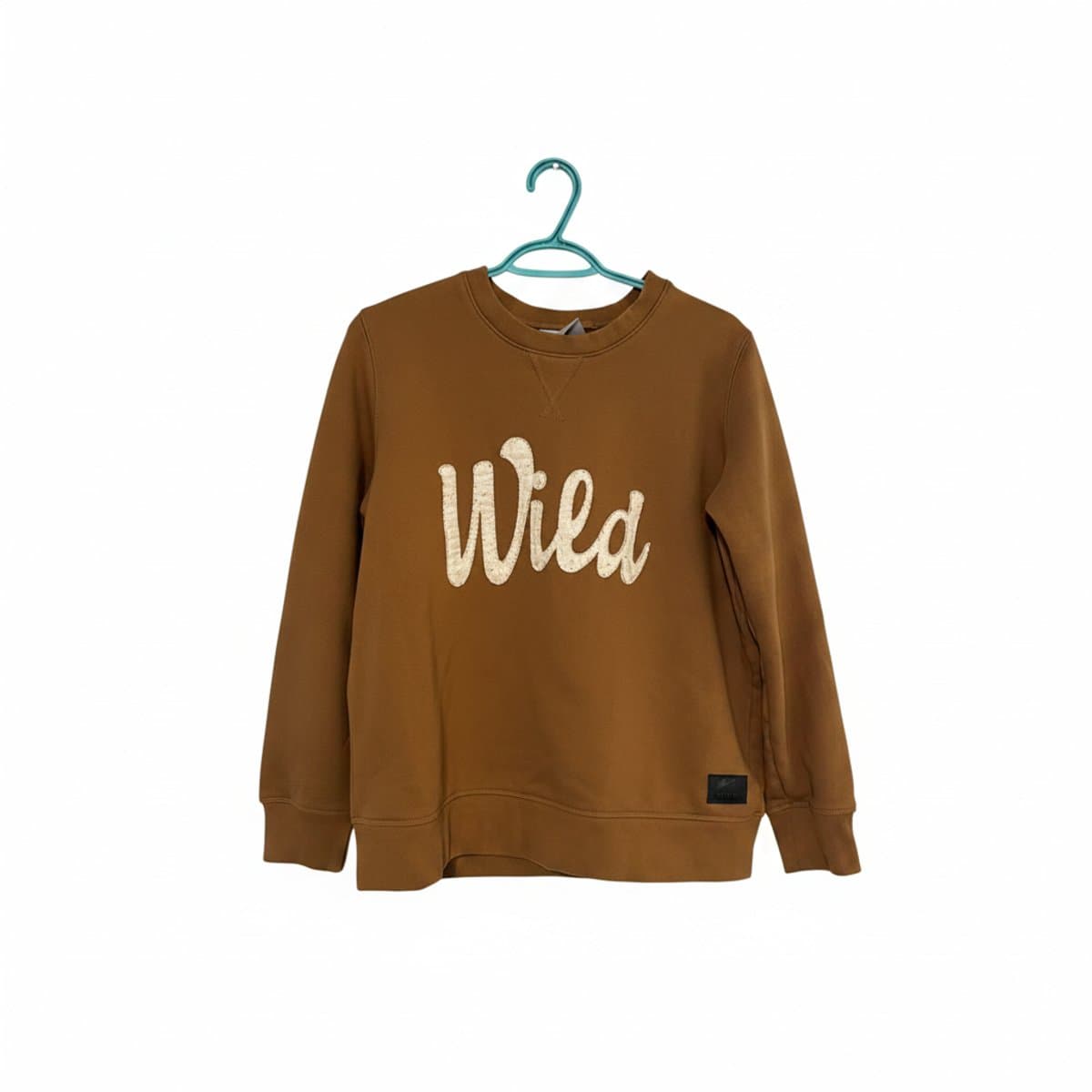 This premium Wild sweater from handpicked306 features a rich, warm mocha-brown hue with a smooth, high-quality cotton-blend jersey texture. The center is adorned with the word "Wild" in a contrasting cream-colored, textured fuzzy chenille appliqué, adding a tactile element to the design. The silhouette is a classic crew-neck pullover with long sleeves, ribbed cuffs, and a matching ribbed hem for a comfortable, structured fit. A subtle v-stitch detail at the collar neckline adds a vintage-inspired touch, while a small, sleek black branded hardware tag is positioned at the lower left hem, completing the sophisticated, minimalist look.
